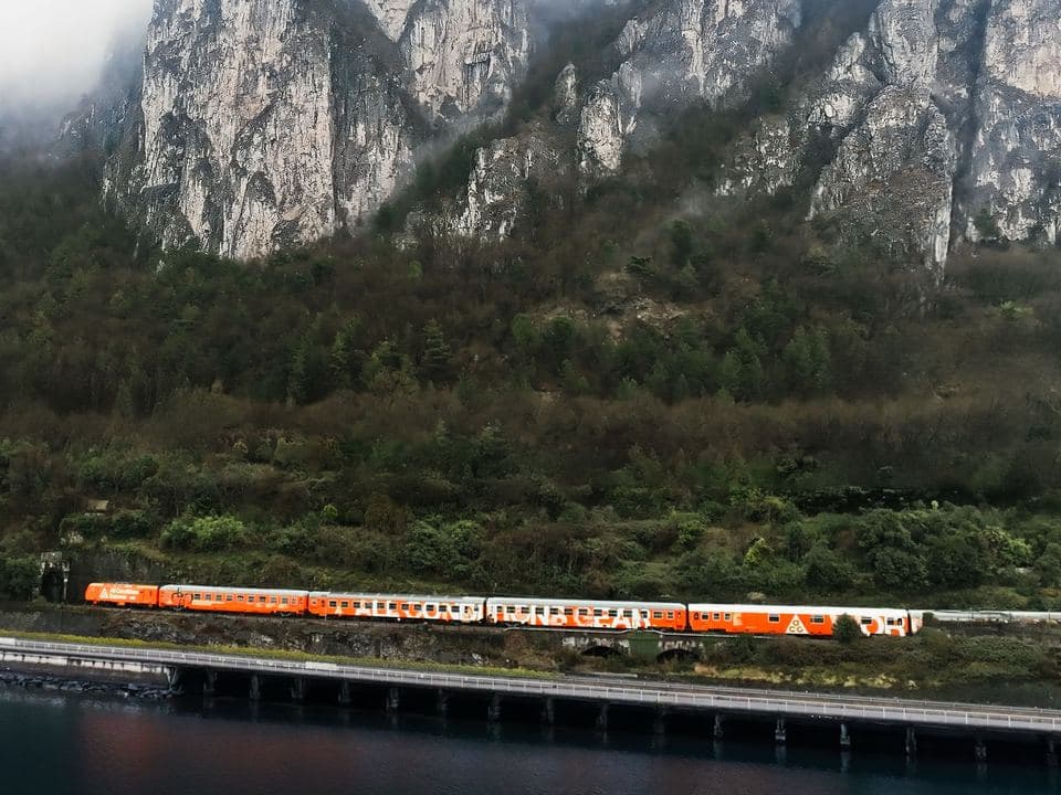 A red train crossing a lakeside under a mountain wall, from ACG’s identity work