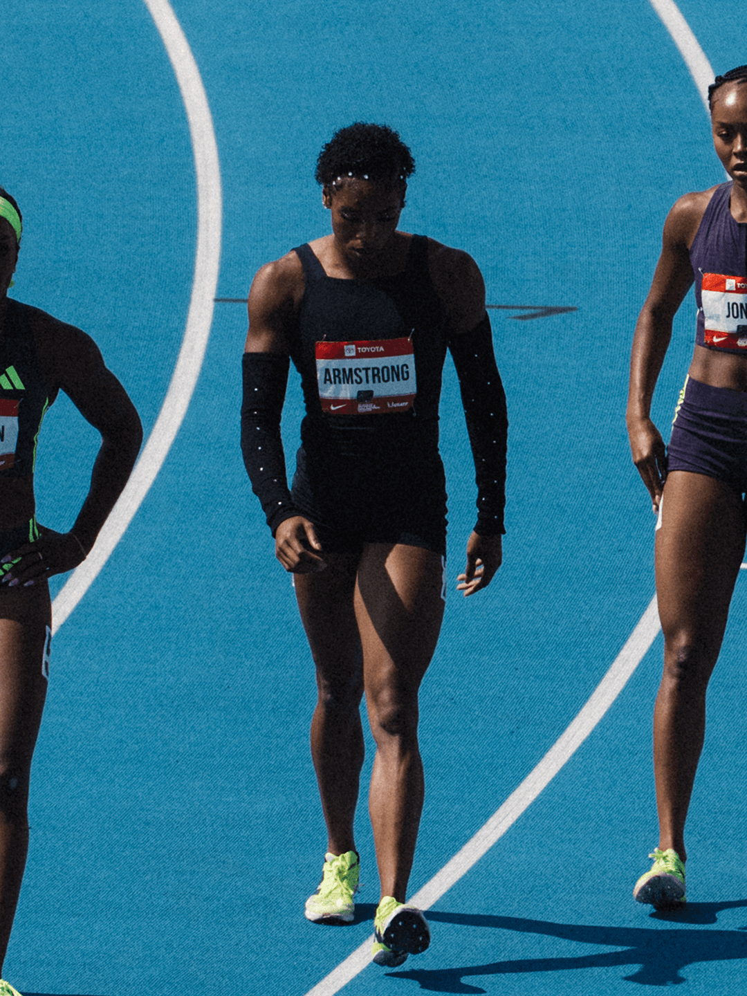 Alia Armstrong walks across Icahn Stadium's blue track in a black long-sleeved one-piece with rhinestones on the sleeves, a competitor in purple behind her