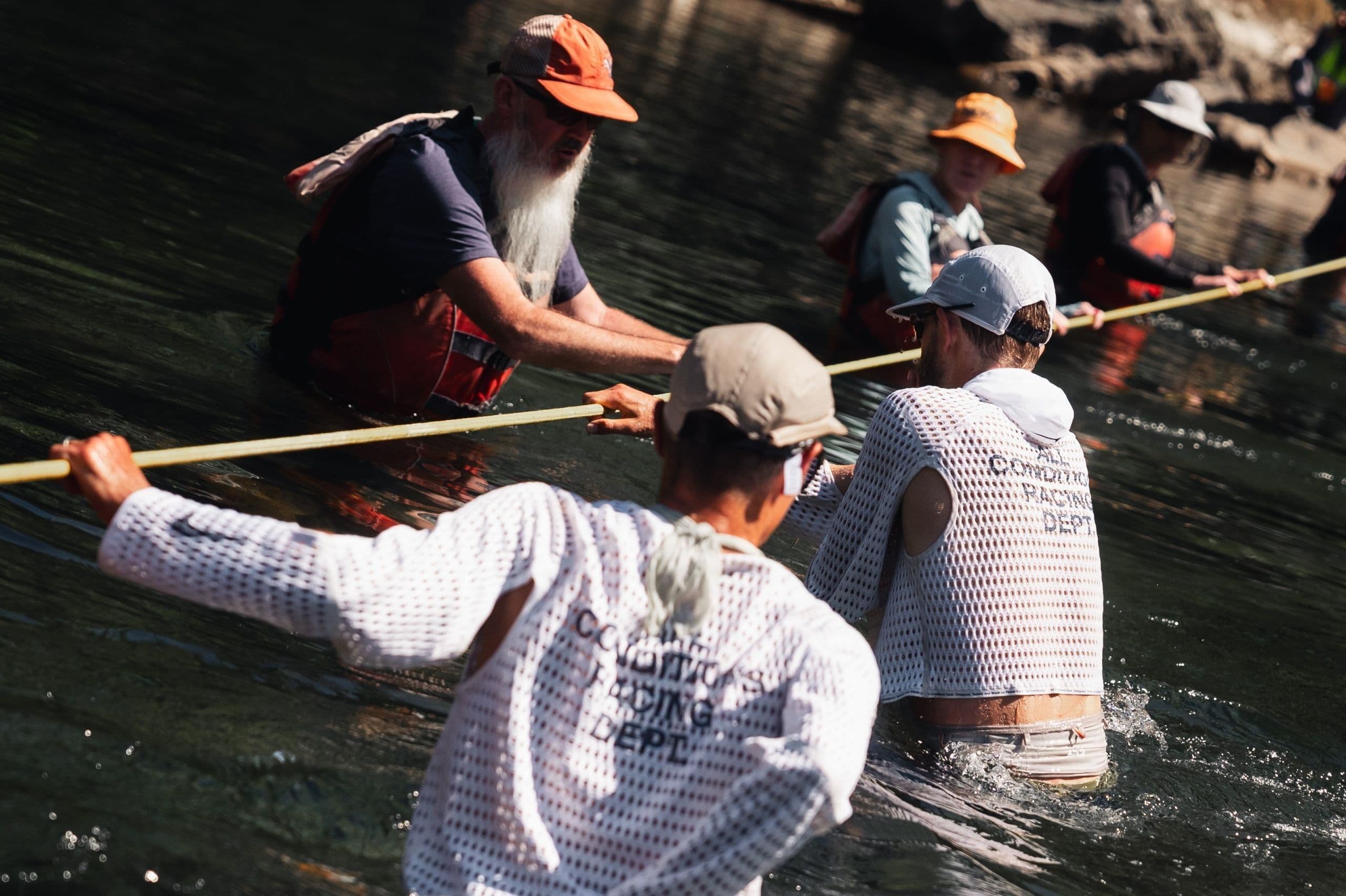 Runners in prototypes crossing a river holding a rope, “All Conditions Racing Dept.” printed on the back