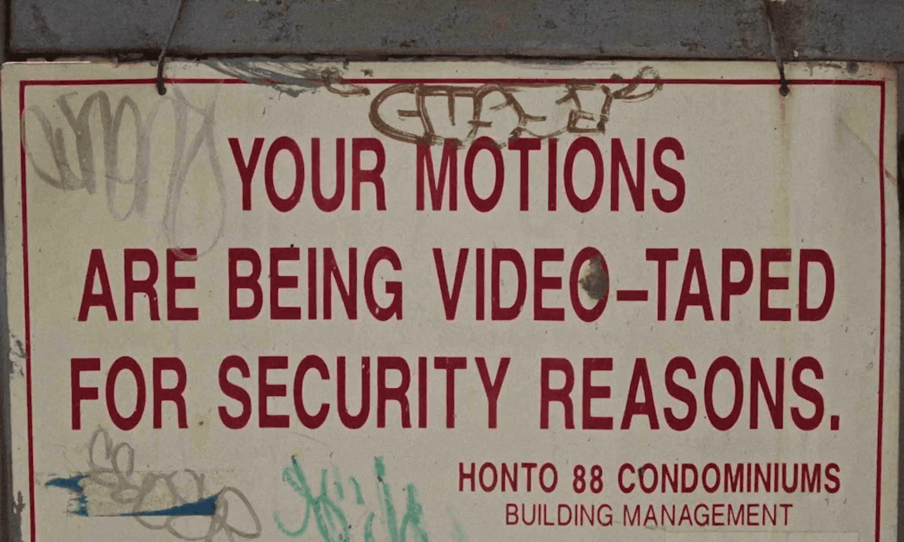 A weathered sign on a condominium wall reading YOUR MOTIONS ARE BEING VIDEO-TAPED FOR SECURITY REASONS, tagged with graffiti