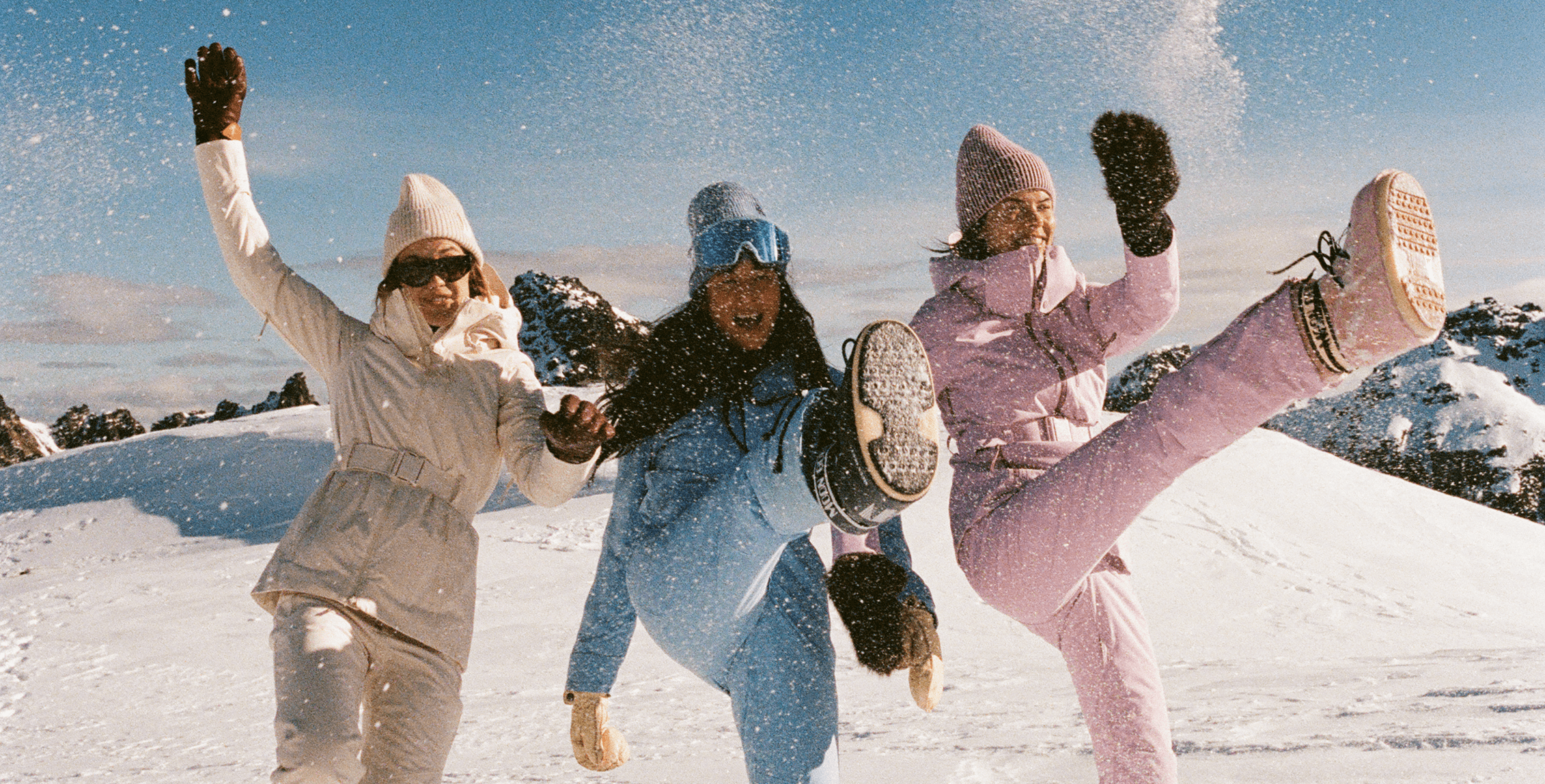 Three women in camel, powder-blue and pink Halfdays ski kit kick up snow on a bluebird day, one mid-air with a boot raised.