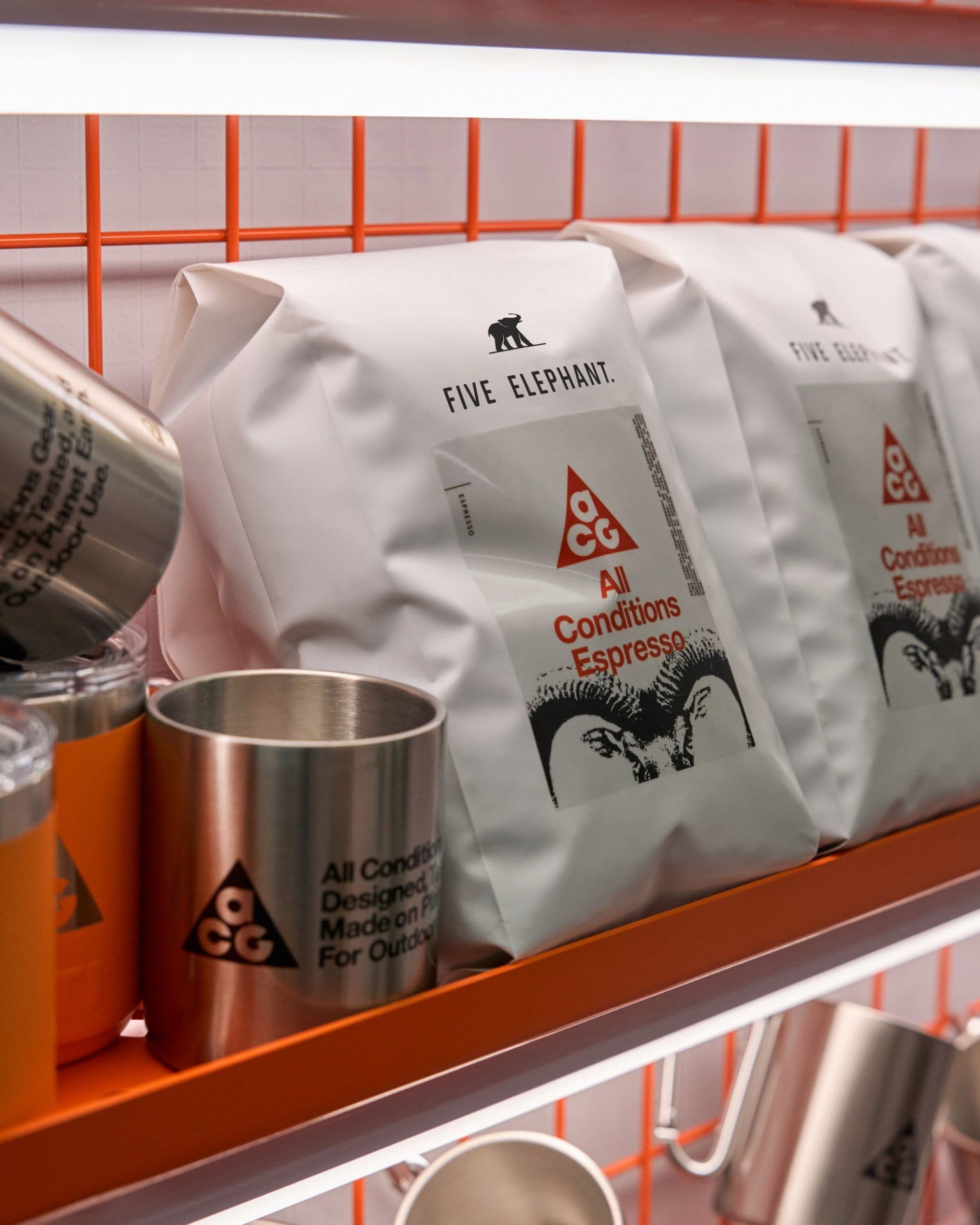 White coffee bags printed All Conditions Espresso with an ibex illustration, beside a steel cup printed All Conditions Gear, on an orange shelf