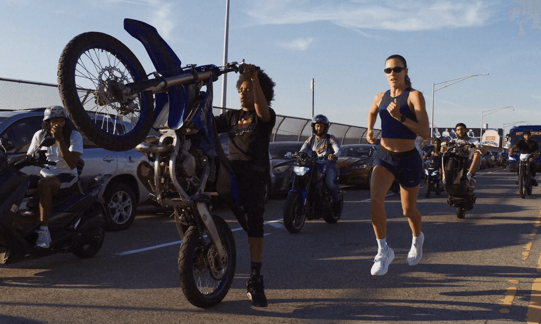 A rider holds a wheelie on a dirt bike beside a woman running in navy kit, a crew of riders behind them in evening light