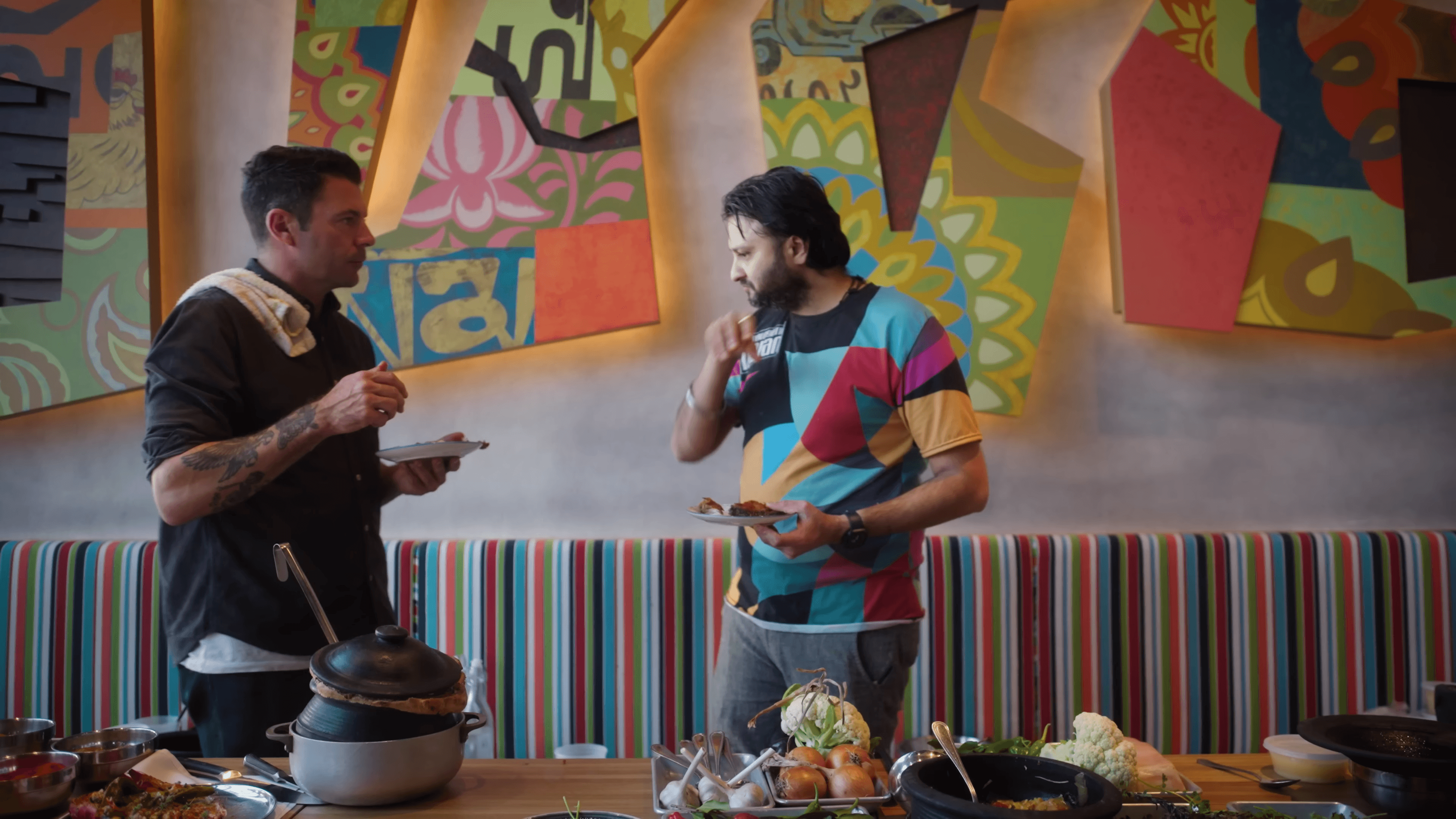 Two men plating food in front of a bright painted mural, a striped banquette and pots on the counter