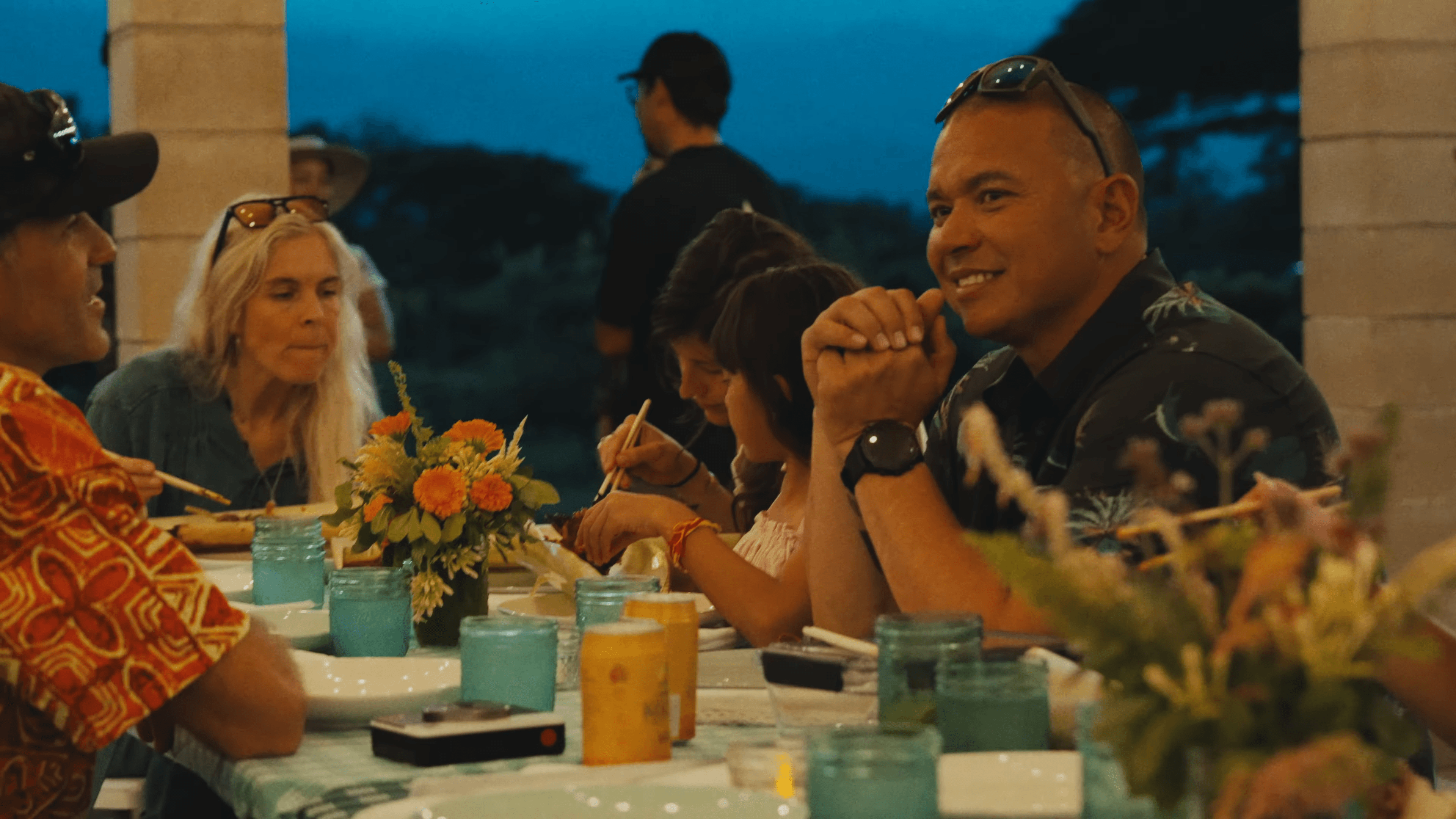 Guests at a long table under a pavilion at blue hour, marigolds and teal glasses down the middle