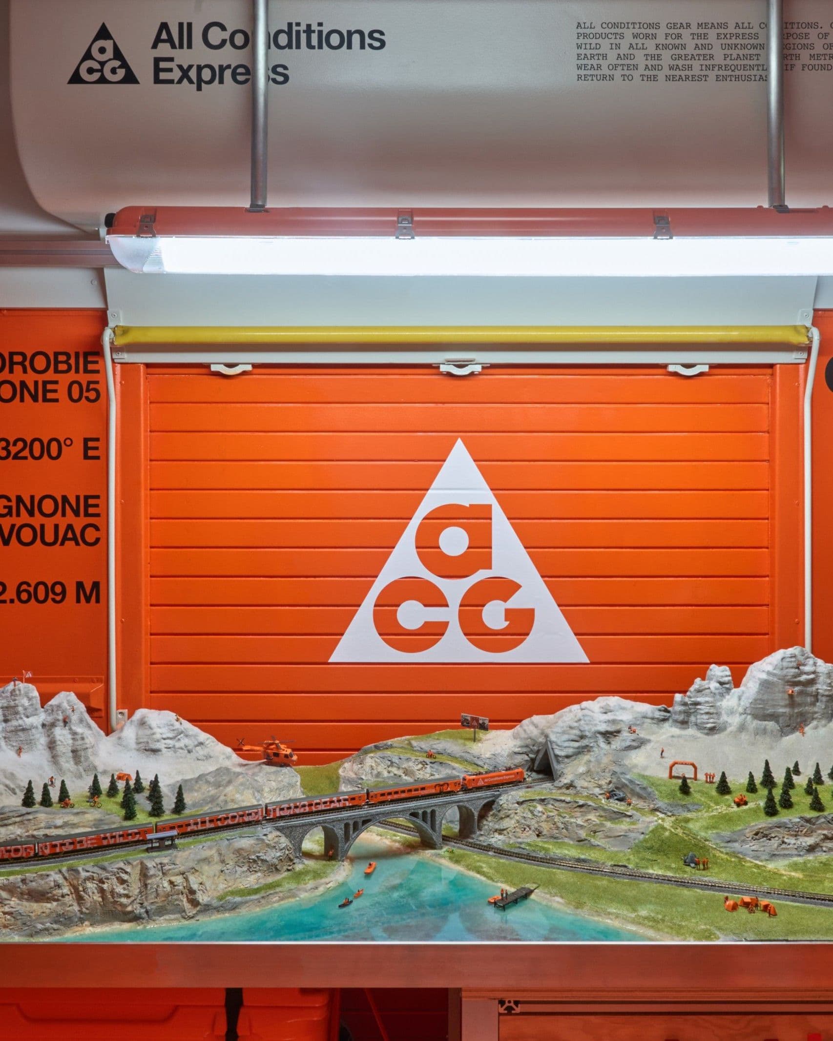 A model railway of the Alpine route under an orange roller shutter printed with a white ACG triangle, with wall type reading ALPI OROBIE GEO ZONE 05 and CITY → WILD