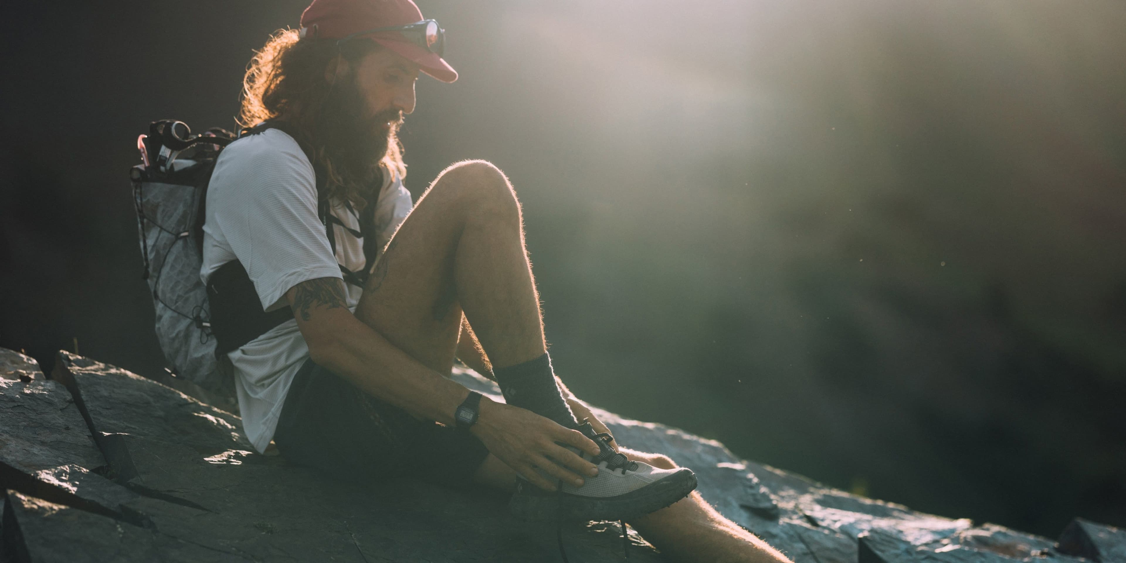 A bearded runner in a red cap sits on a rock ledge in low sun, lacing a pale grey trail shoe, a white pack on his back