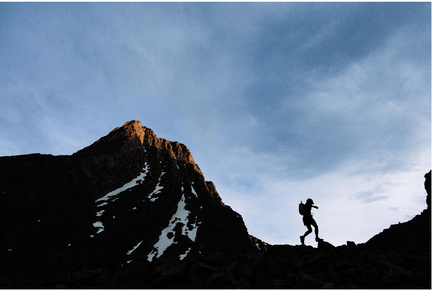 A runner in silhouette crosses a dark ridge toward a peak lit orange at its summit, snow in the gullies