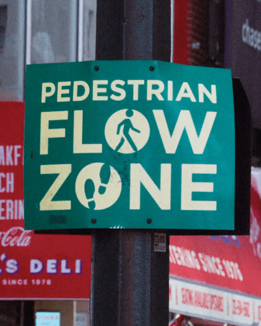 A green street sign on a pole reading PEDESTRIAN FLOW ZONE, with a walking figure in the O of FLOW and a shoe print in the O of ZONE, deli awnings behind