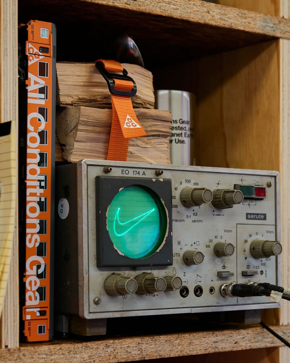 A vintage oscilloscope tracing a green Swoosh on its screen, beside an orange spine lettered All Conditions Gear