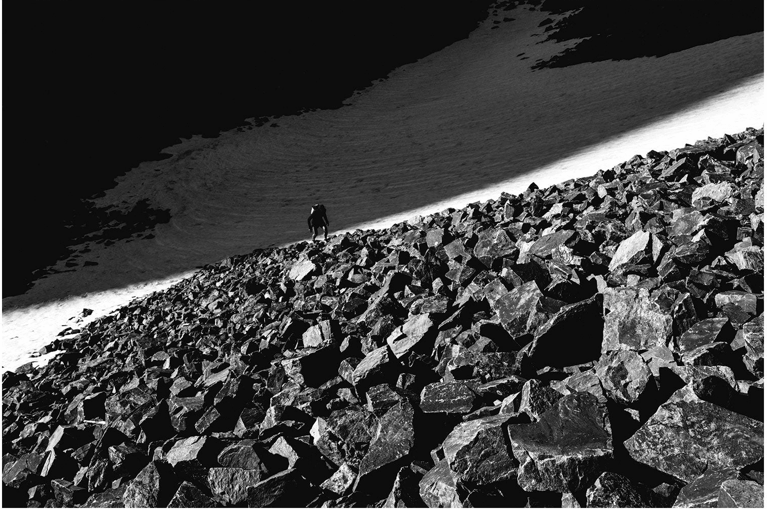 Black and white: a lone figure on a steep talus field below a sloping band of snow