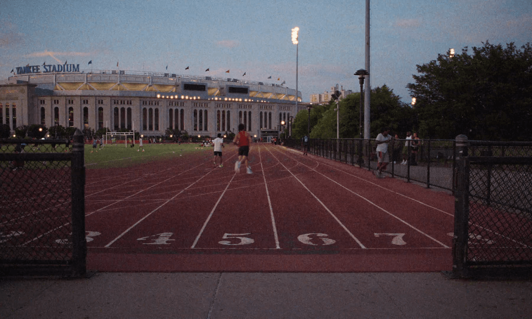 A running track at dusk with Yankee Stadium lit up behind it, two runners small in the far lanes