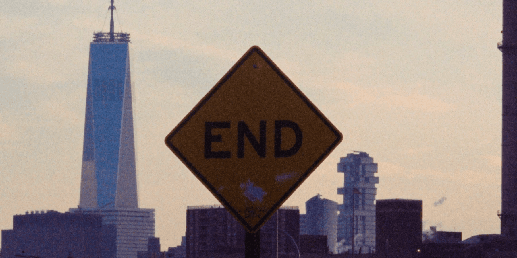 A yellow diamond road sign reading END in the foreground, One World Trade Center rising behind it against a dusk sky