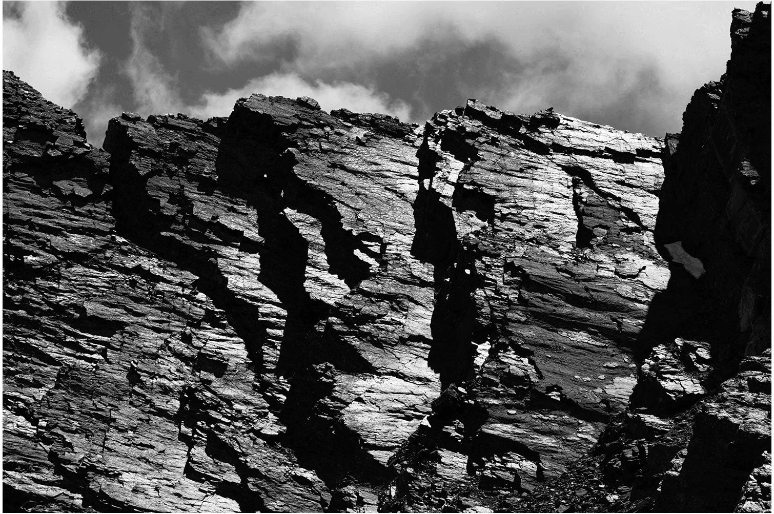 Black and white: a wall of layered, fractured rock under cloud