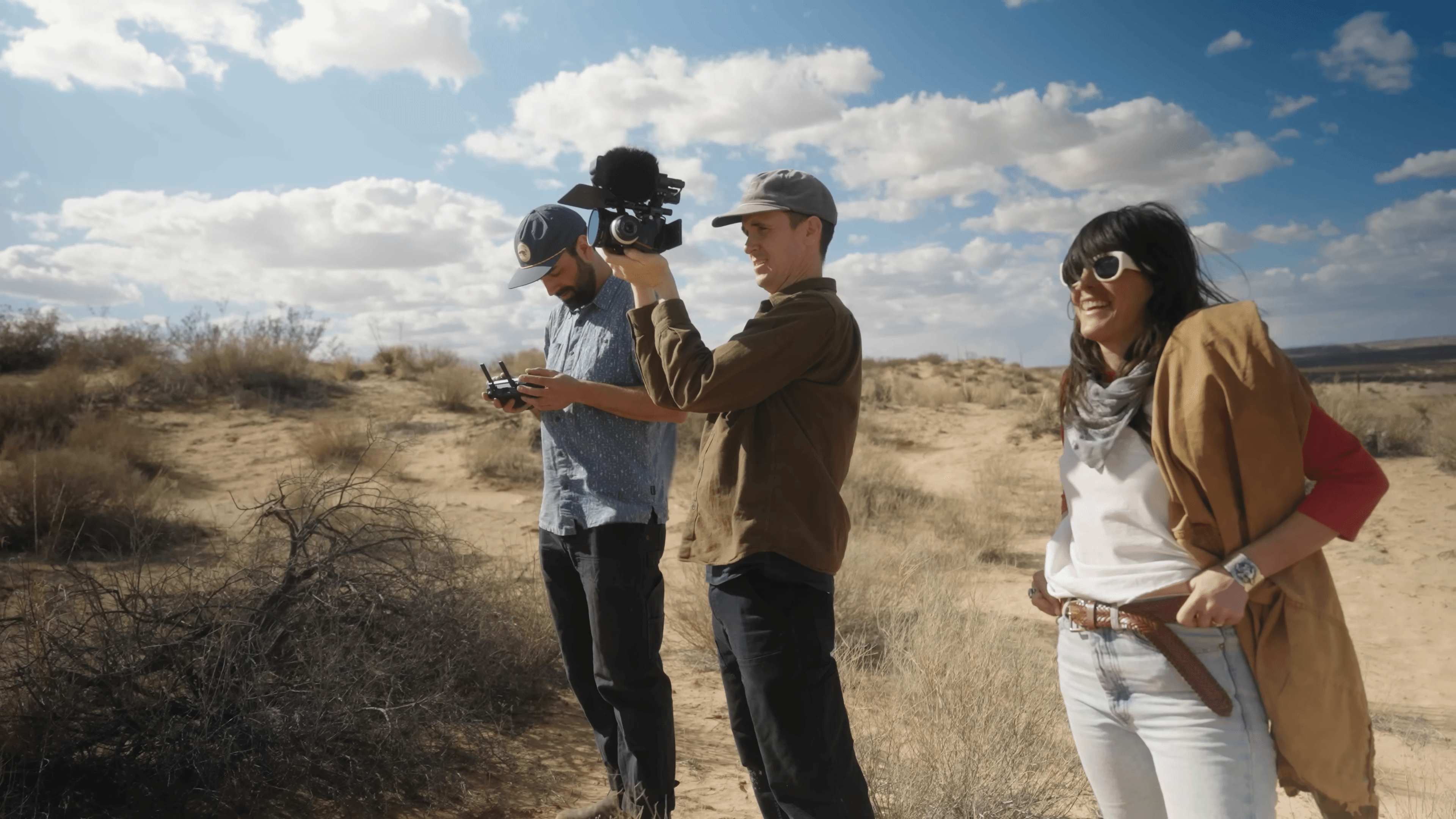 Two camera operators with a drone controller and a cinema camera in high desert scrub, Kate West beside them in sunglasses