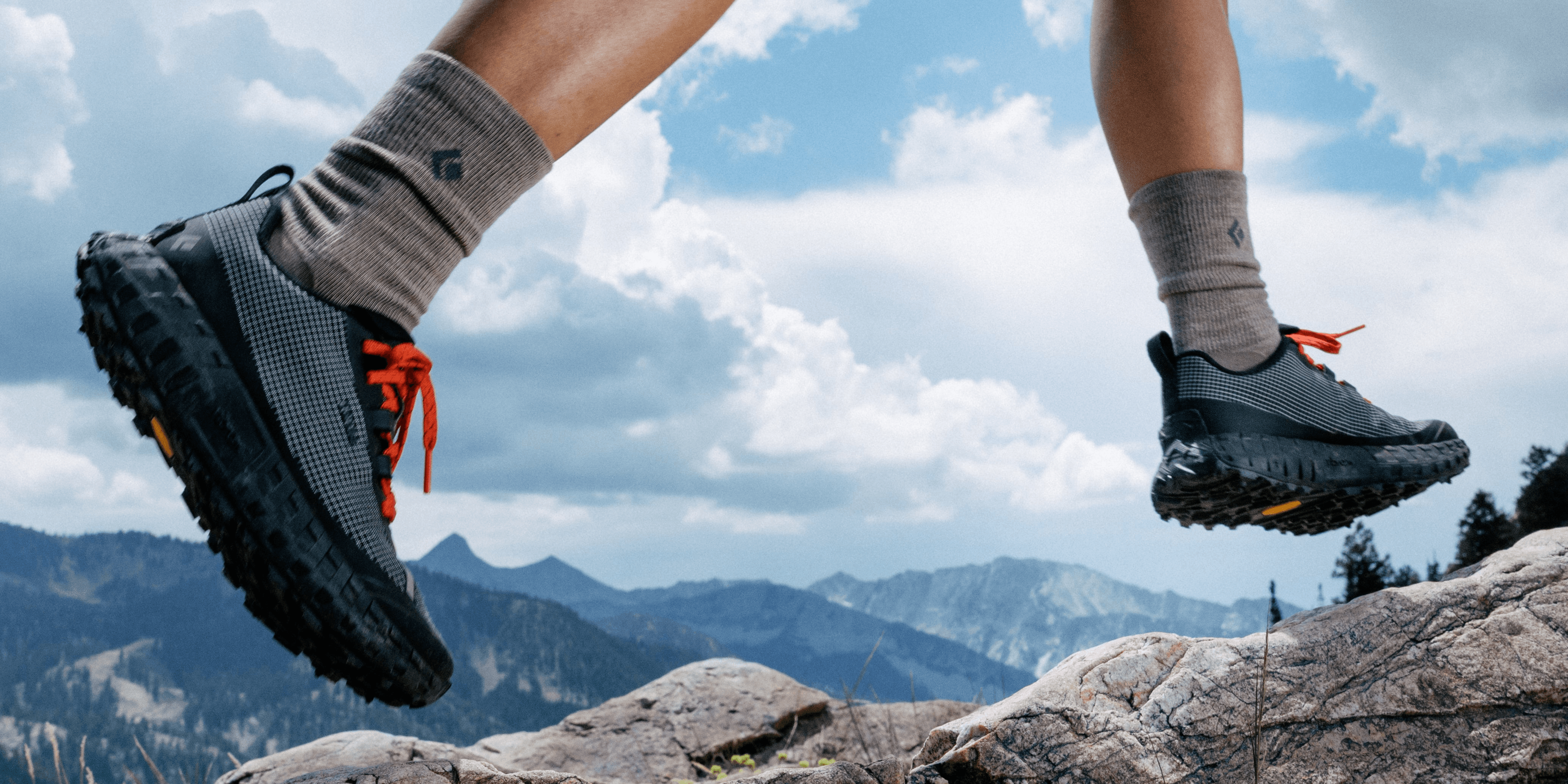 A runner's legs mid-stride over pale rock in two black and grey trail shoes with red laces, mountains and cloud behind