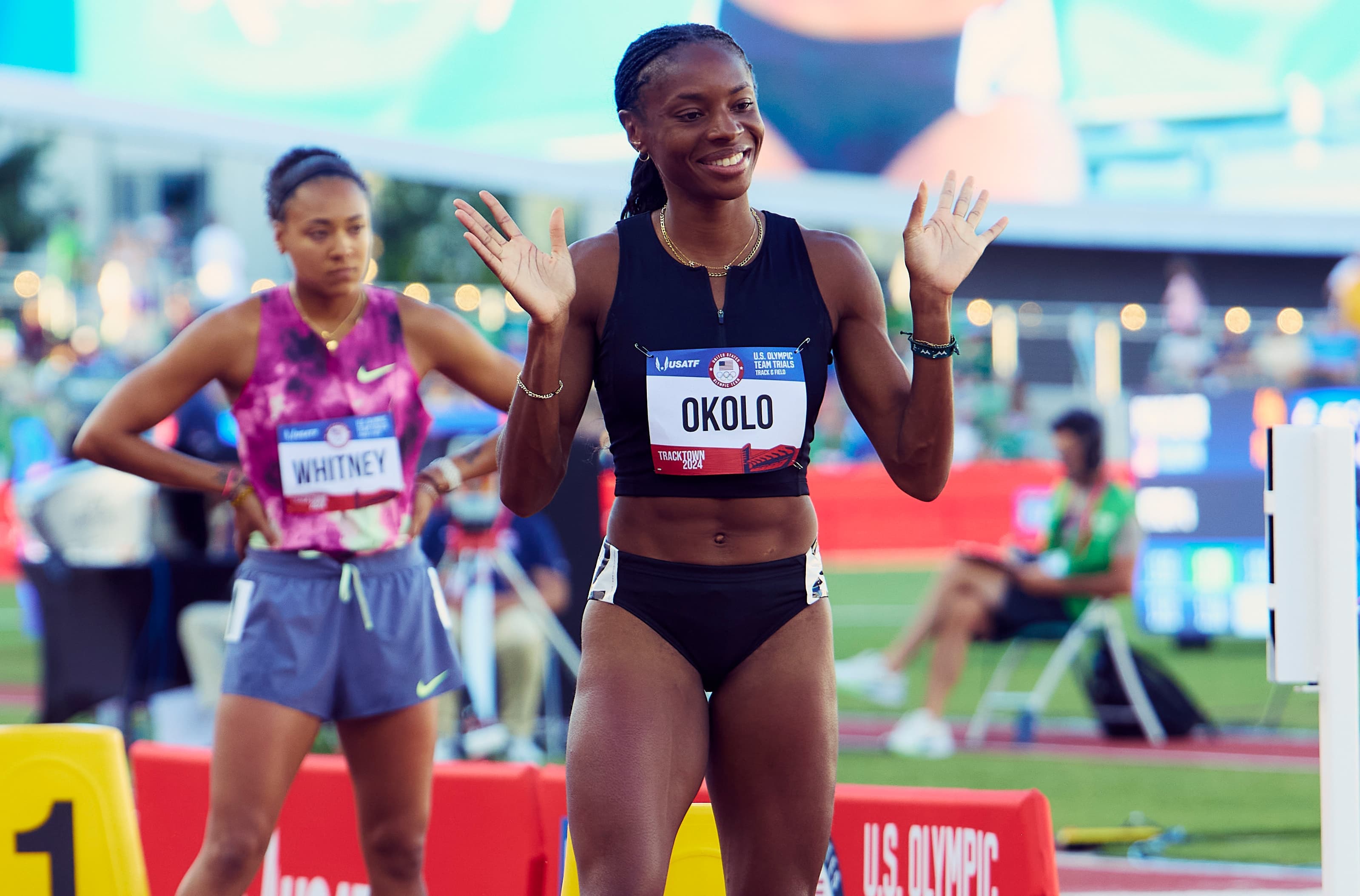 Courtney Okolo smiles and waves at the 2024 Olympic Trials in a plain black crop top and briefs, beside a competitor in a Nike tie-dye top