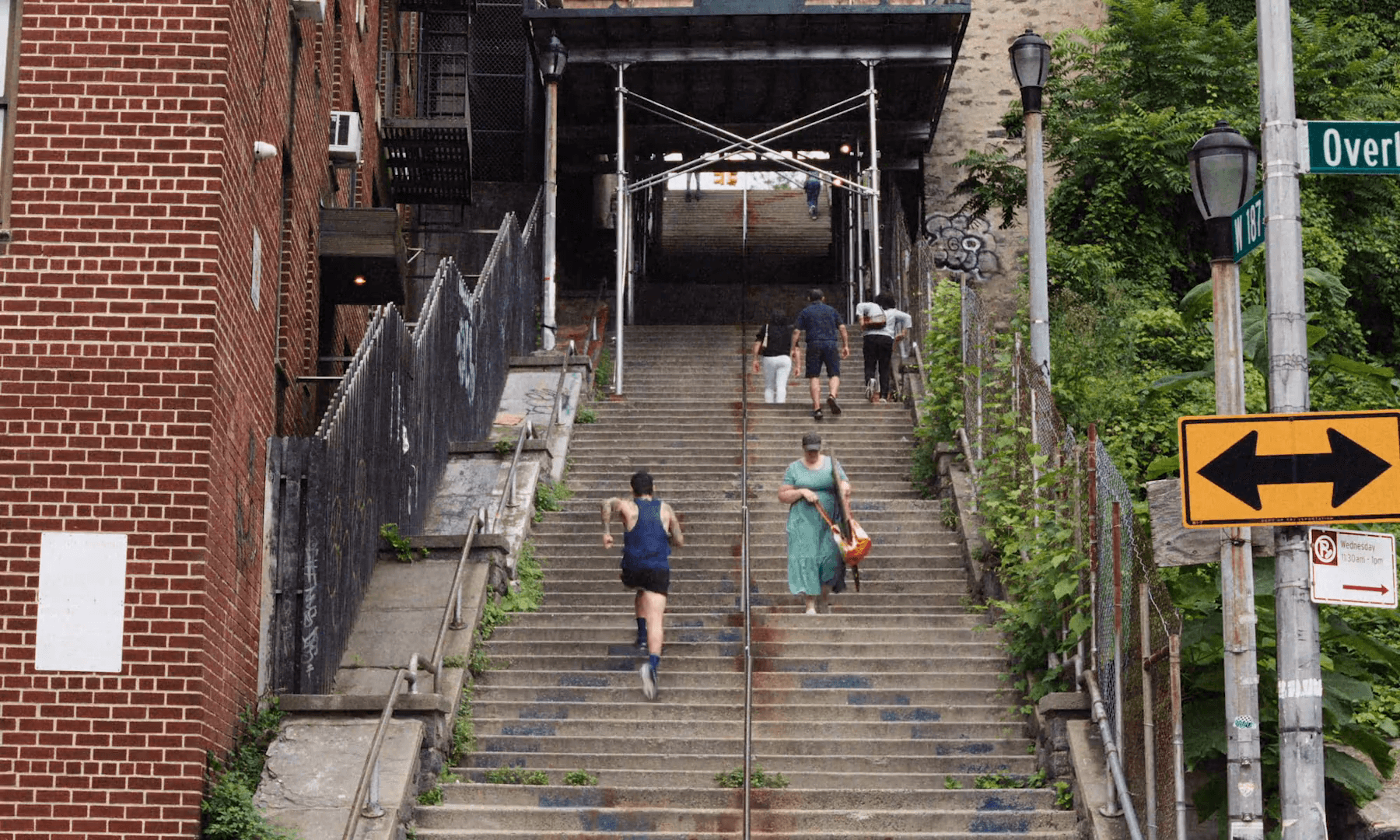 A runner in a navy singlet climbs the West 187th Street step street between brick walls and a green street sign
