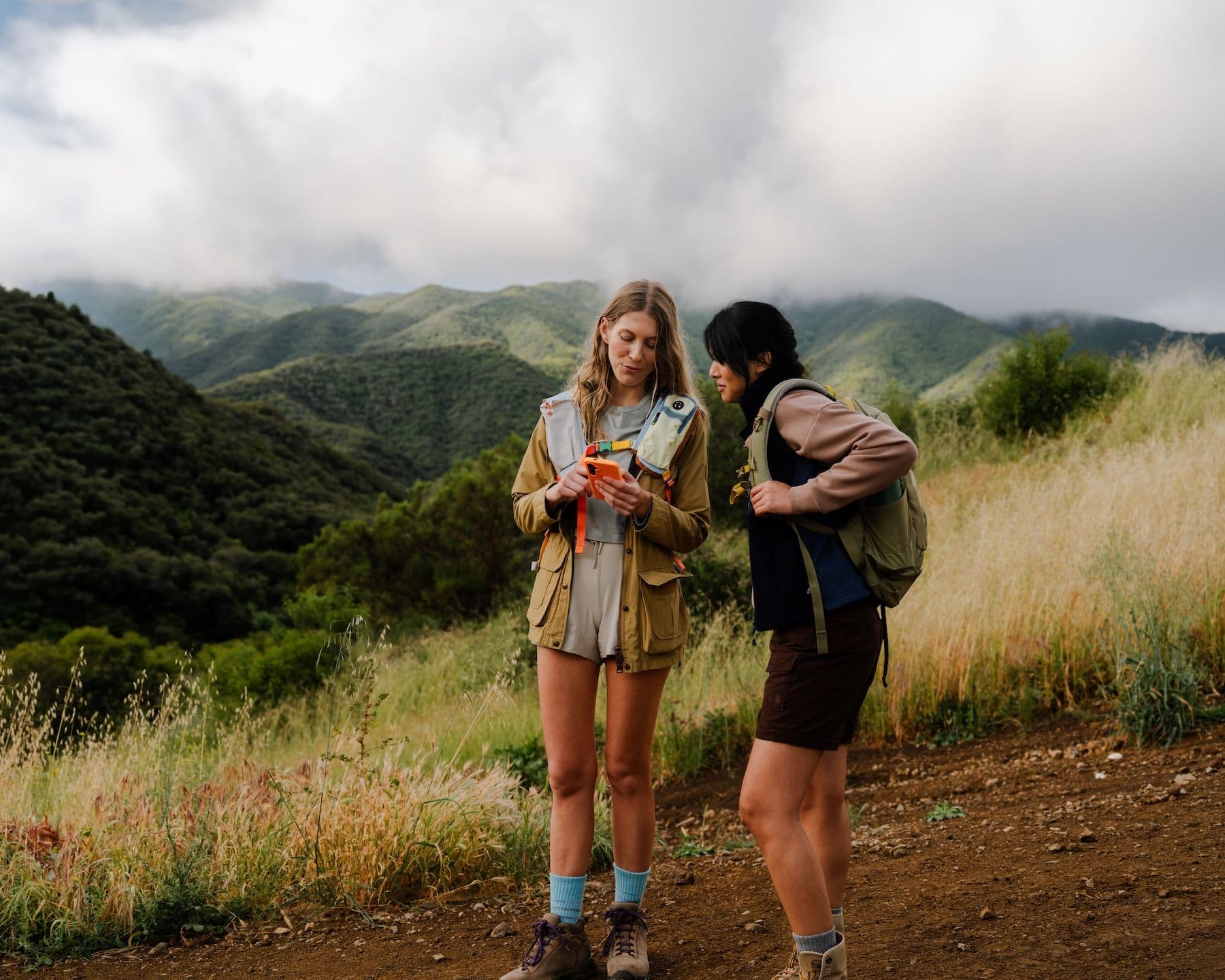 Two women on a dirt trail on a grassy ridge, green hills and low cloud behind, both looking at a phone one of them is holding.