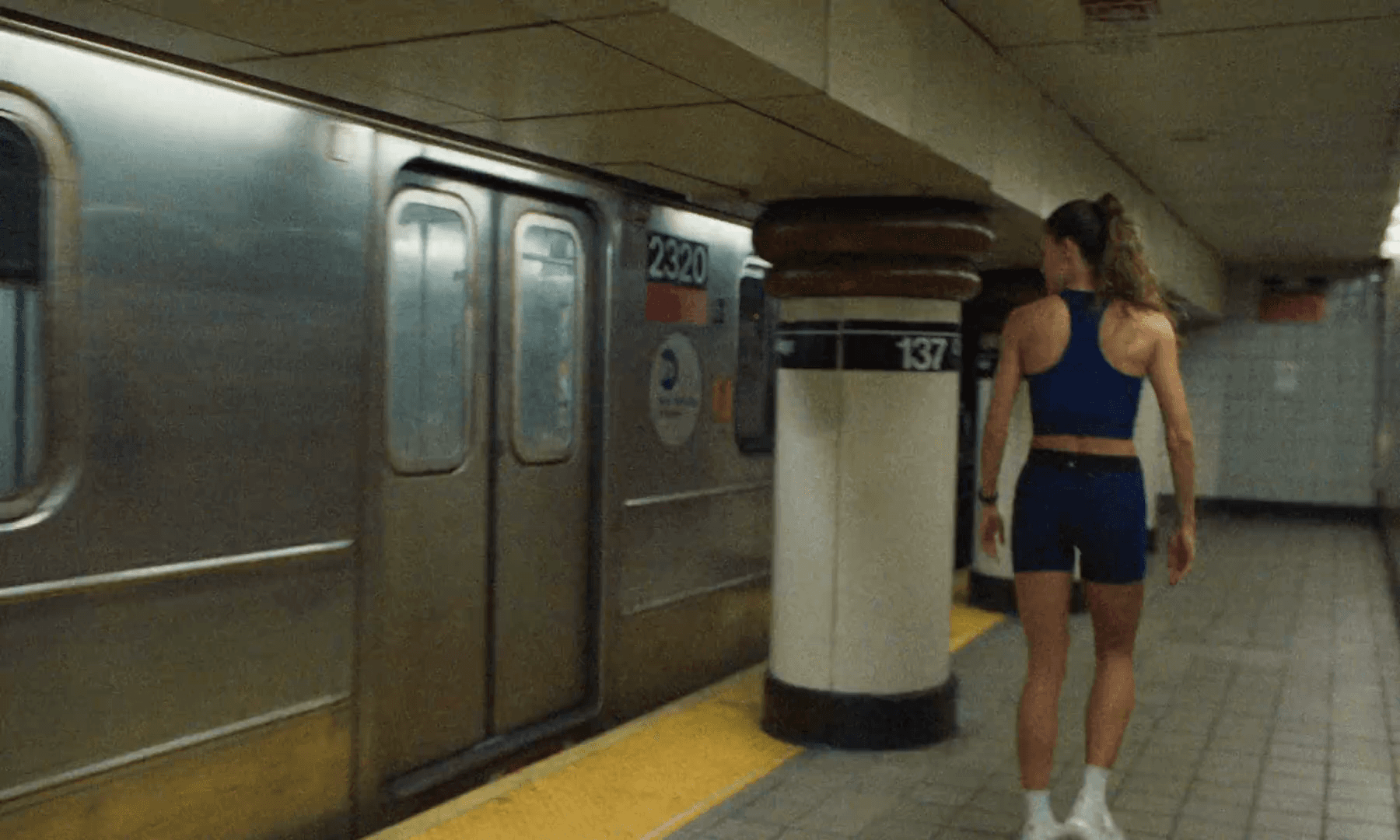 A woman in a navy run bra and shorts walks a subway platform beside a standing 1 train at 137th Street