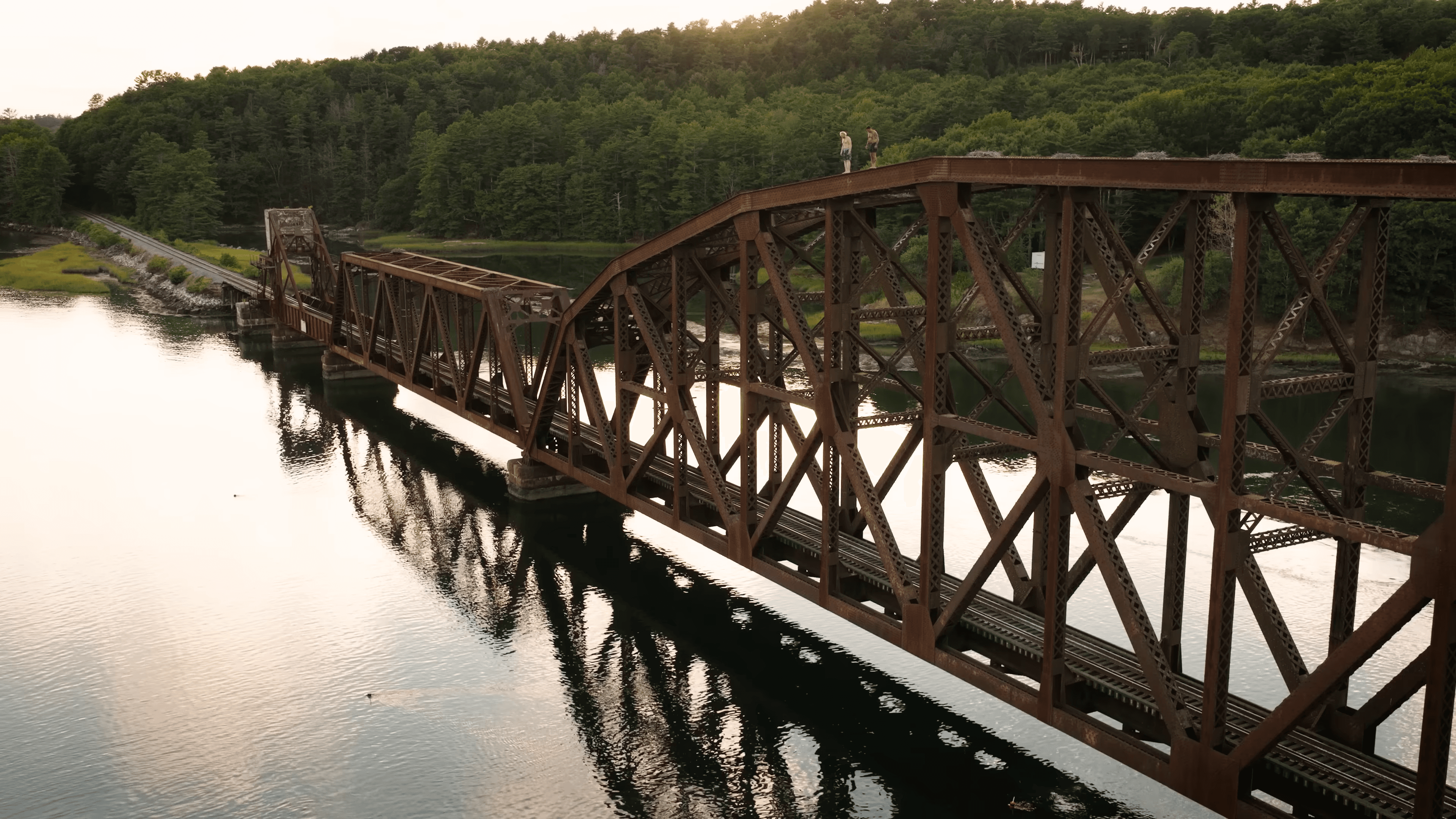 An aerial view down a rusted steel rail trestle over still water at golden hour, two men standing on its top chord, forest behind