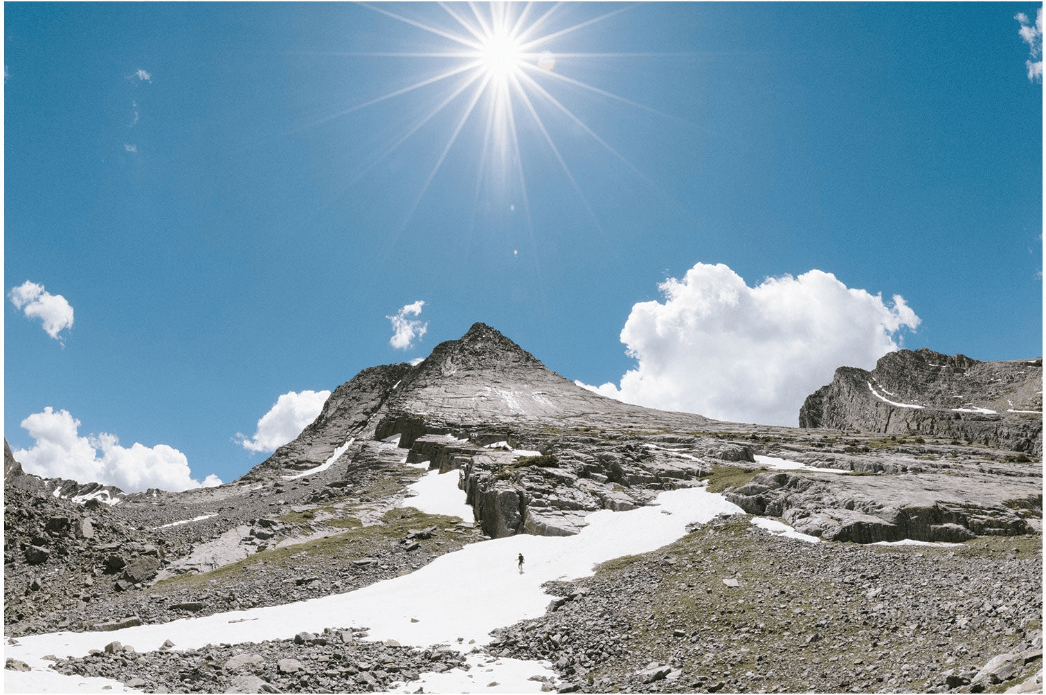 A sunburst over a high basin of grass, snow patches and rock, a tiny figure crossing the snow