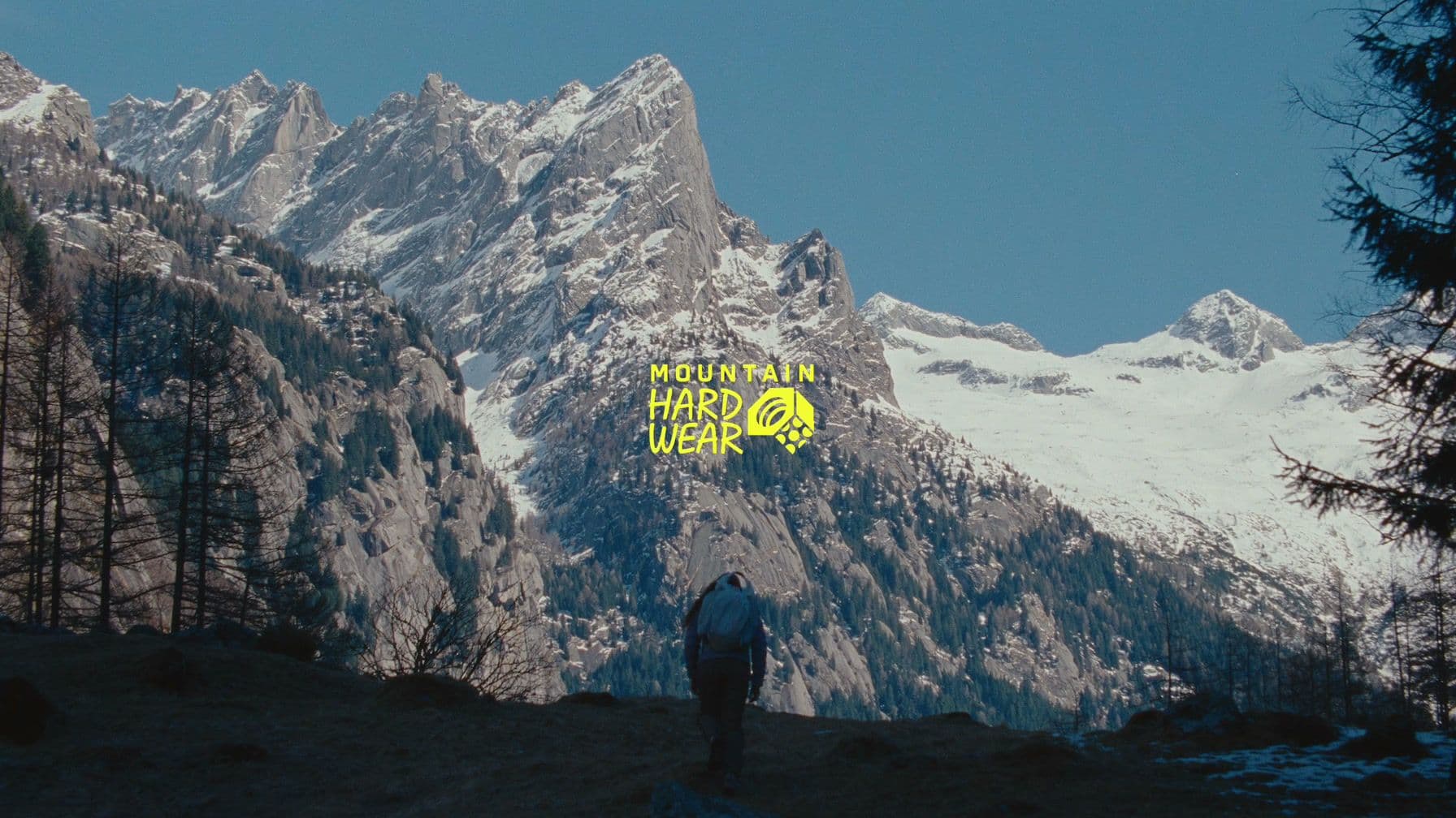 A lone hiker with a backpack walks toward a valley of snow-capped granite peaks, with the Mountain Hardwear wordmark in yellow at center frame.