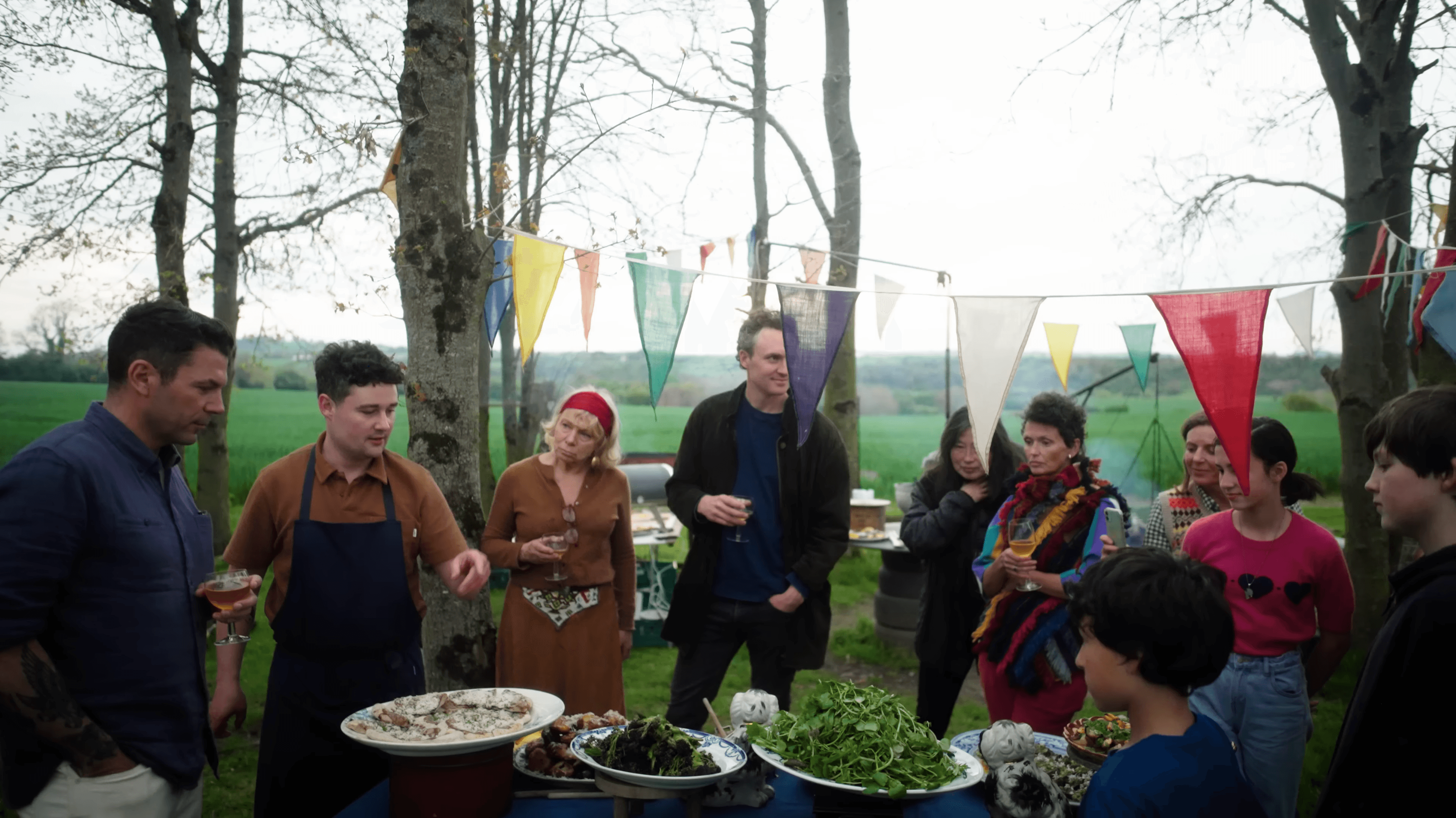 A crowd gathered around platters of seafood and greens under strings of coloured bunting in a field
