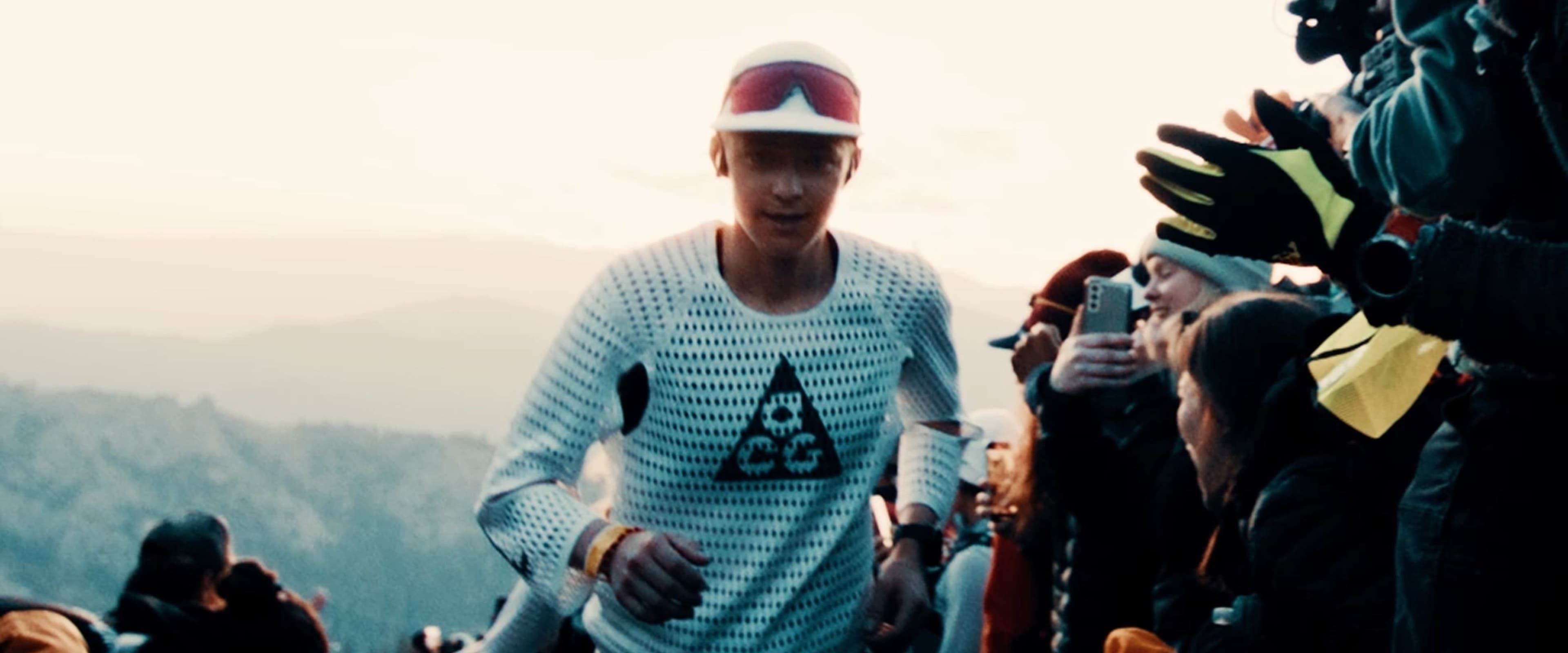 Caleb Olson runs up the Escarpment at dawn in a white perforated long-sleeve with the ACG triangle, spectators reaching in with phones