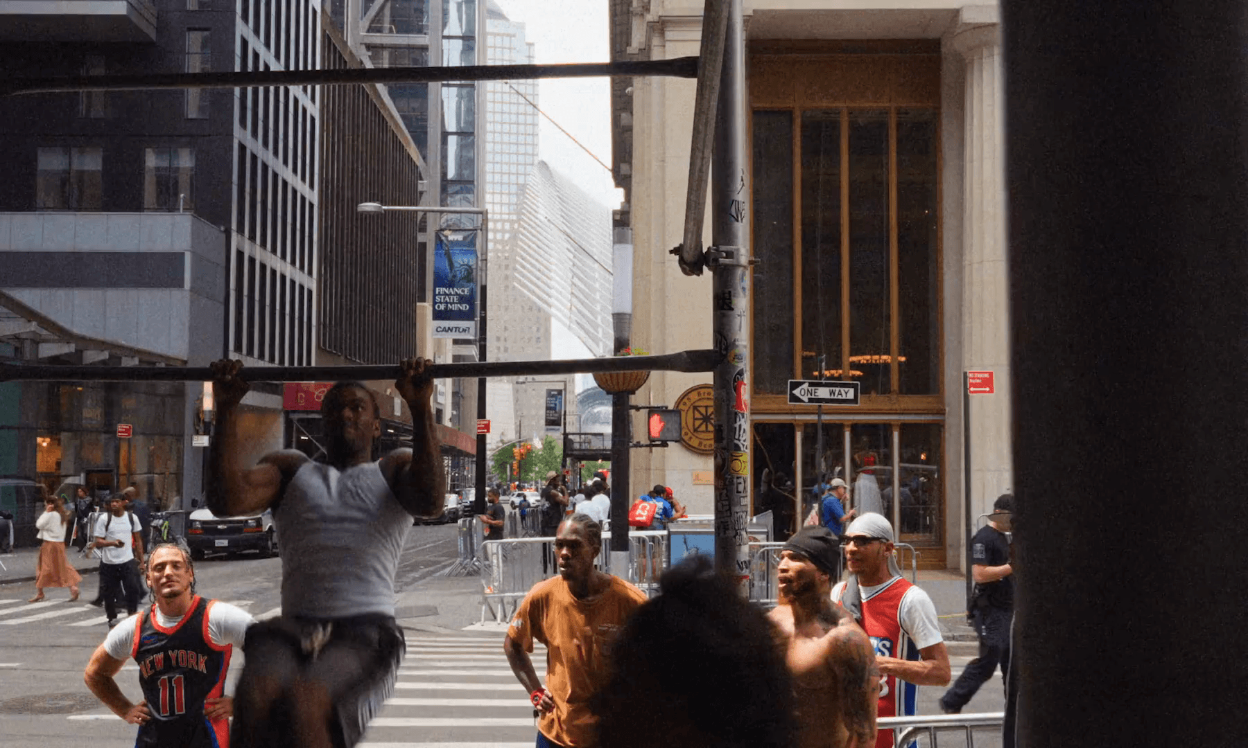 A man does pull-ups on a scaffold bar in the Financial District, the Oculus visible down the street, onlookers around him