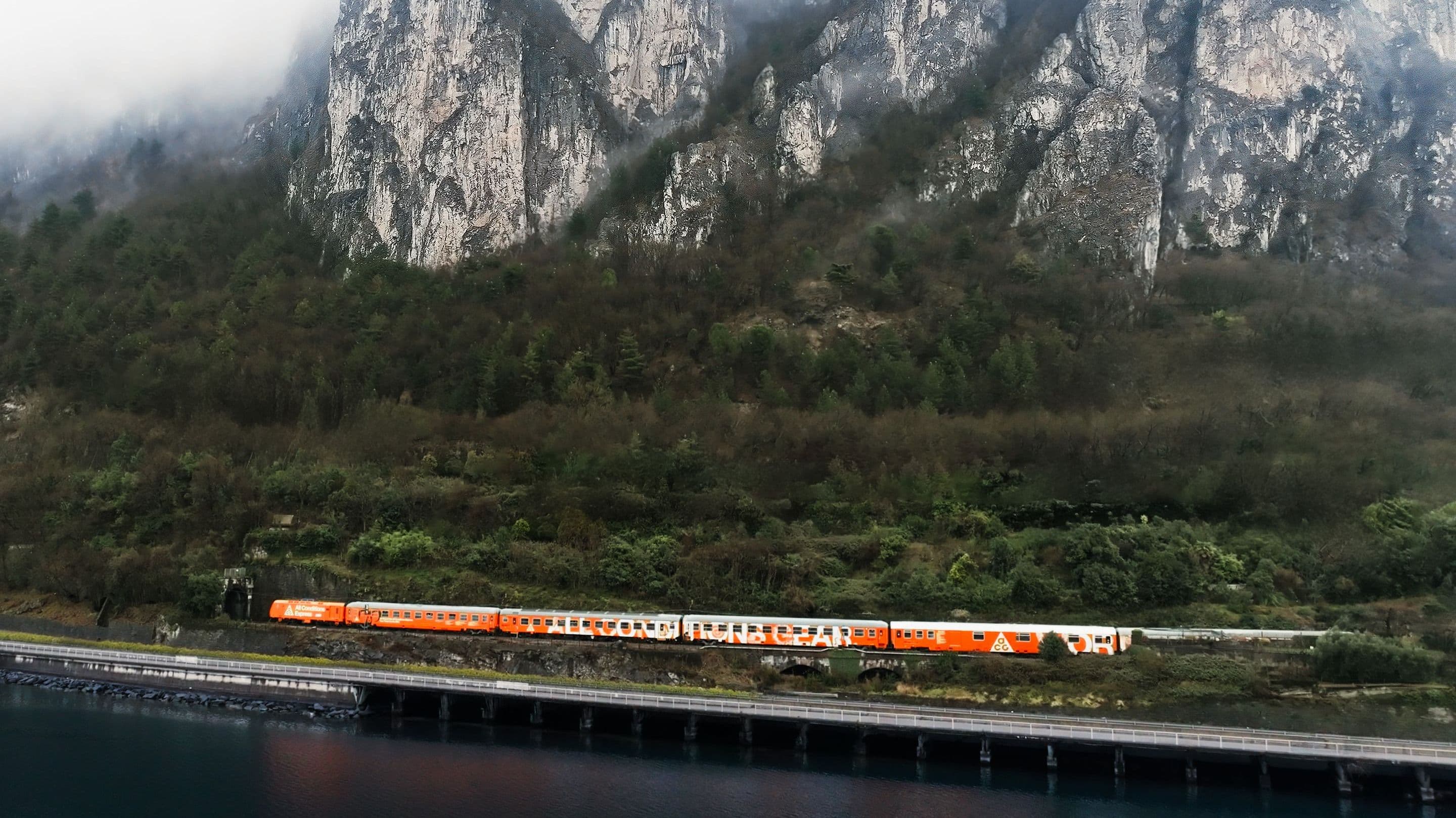 The All Conditions Express, a thin orange line of carriages, running along a lakeshore beneath mist-capped limestone cliffs