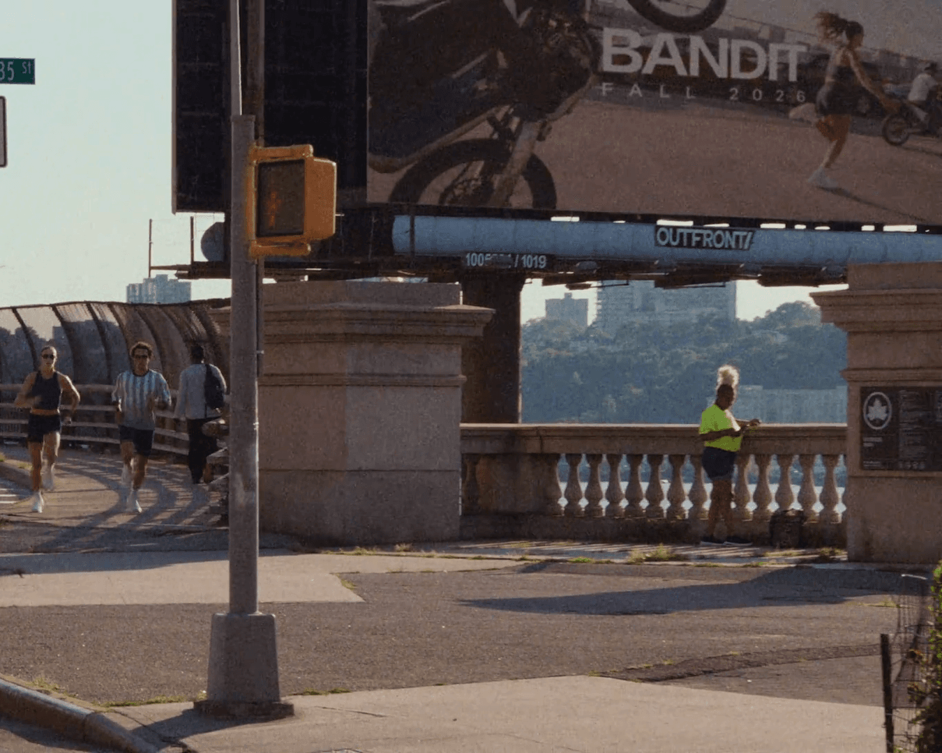 Two runners come off the Riverside Drive viaduct at West 135th Street beneath an Outfront billboard reading BANDIT FALL 2026