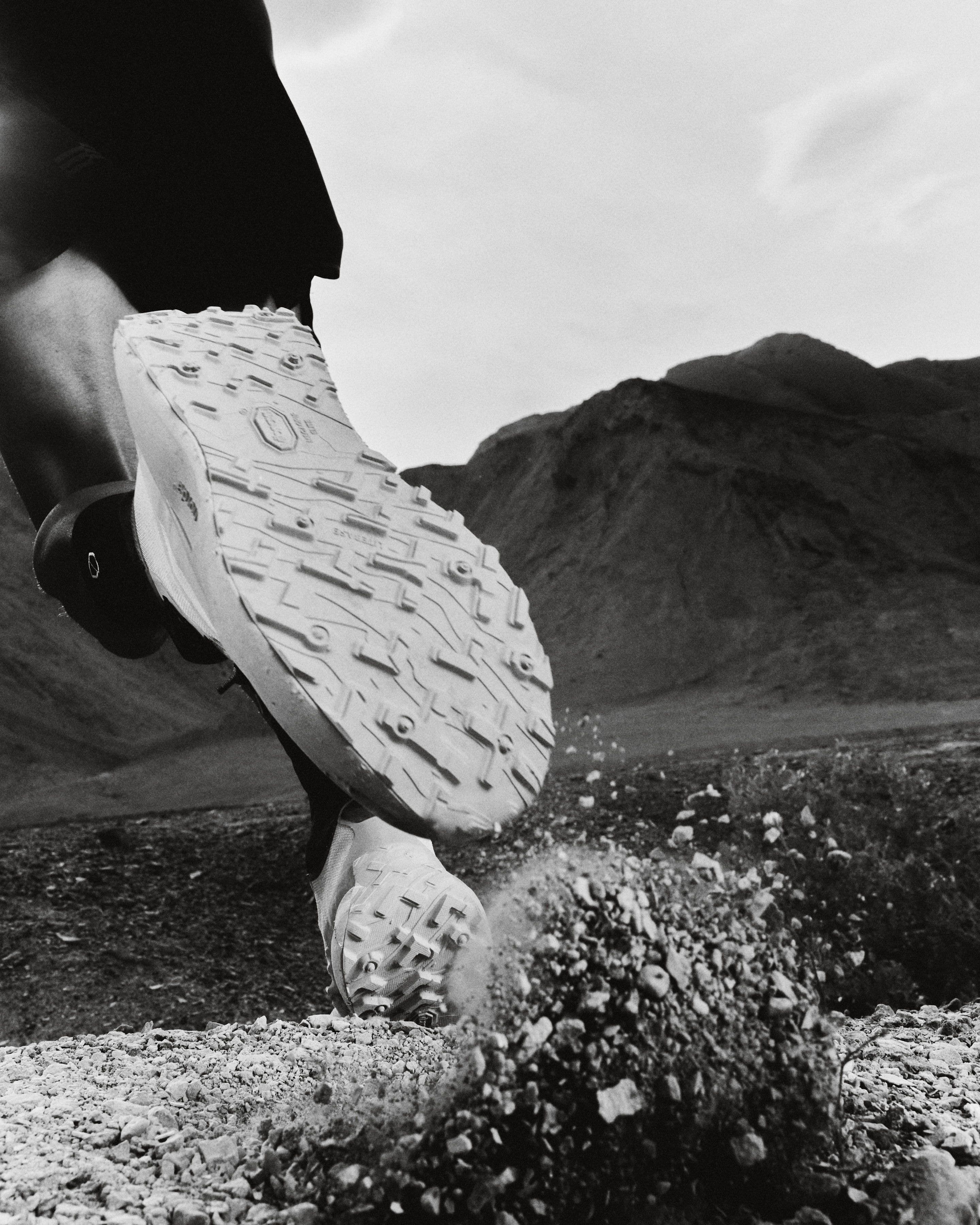 An outsole lifted mid-stride, lug pattern to camera, a desert ridge behind