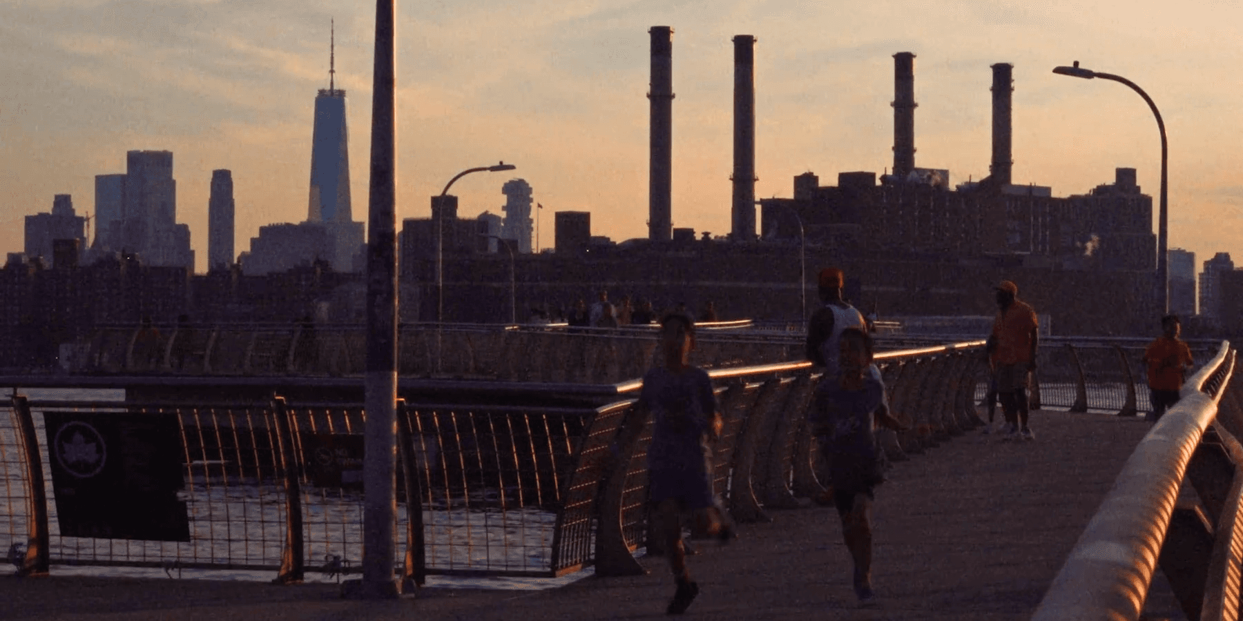 Golden hour on a waterfront promenade; two children sprint toward the camera past runners, with One World Trade and four power-station smokestacks on the skyline behind
