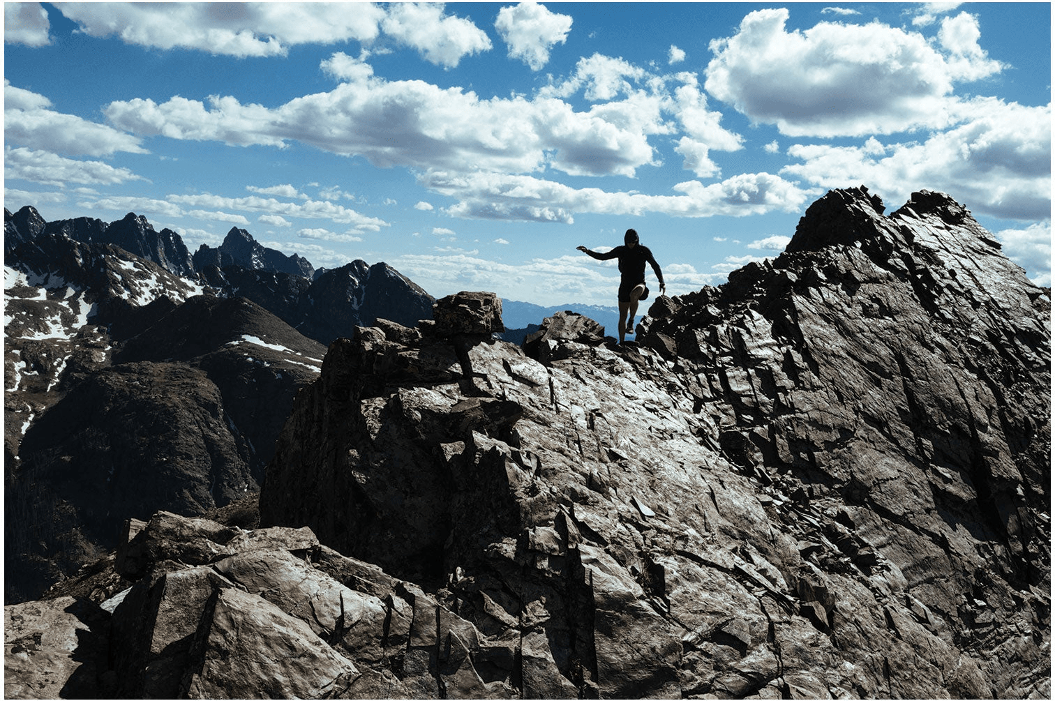 A runner stands on a jagged rock summit under a blue sky full of cloud, serrated peaks behind