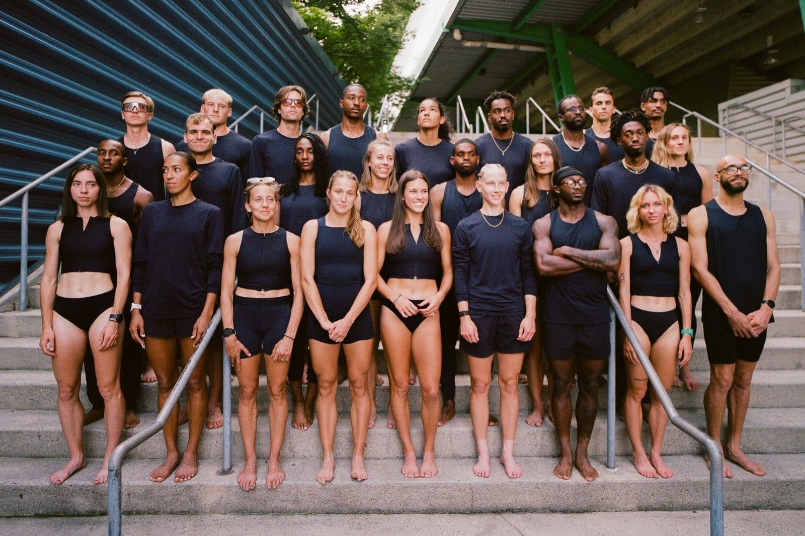 About twenty-eight athletes standing barefoot on concrete stadium stairs under green steel stands, all in the same near-black crops, tanks and long-sleeves with no logo anywhere